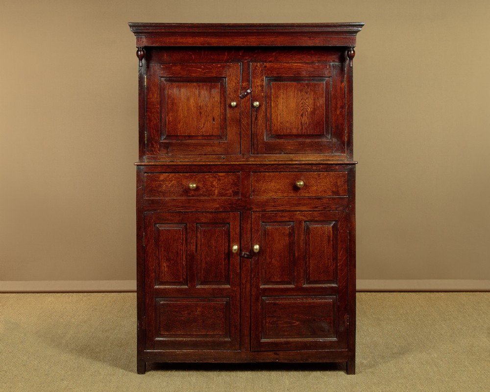 An 18th-century dark Welsh oak cwpwrdd deuddarn featuring an overhanging canopy with drop pendants, upper fielded panel doors, two drawers, and lower cupboard doors. View II