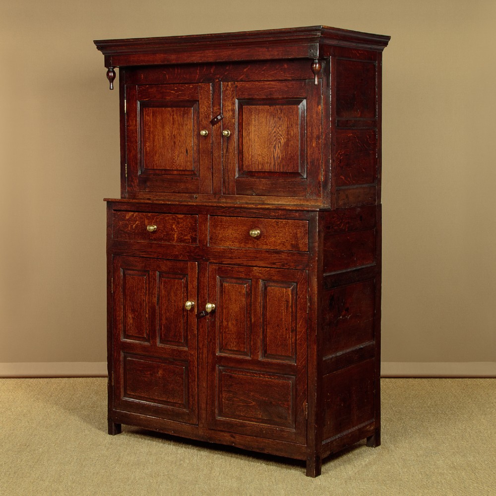 An 18th-century dark Welsh oak cwpwrdd deuddarn featuring an overhanging canopy with drop pendants, upper fielded panel doors, two drawers, and lower cupboard doors. View III