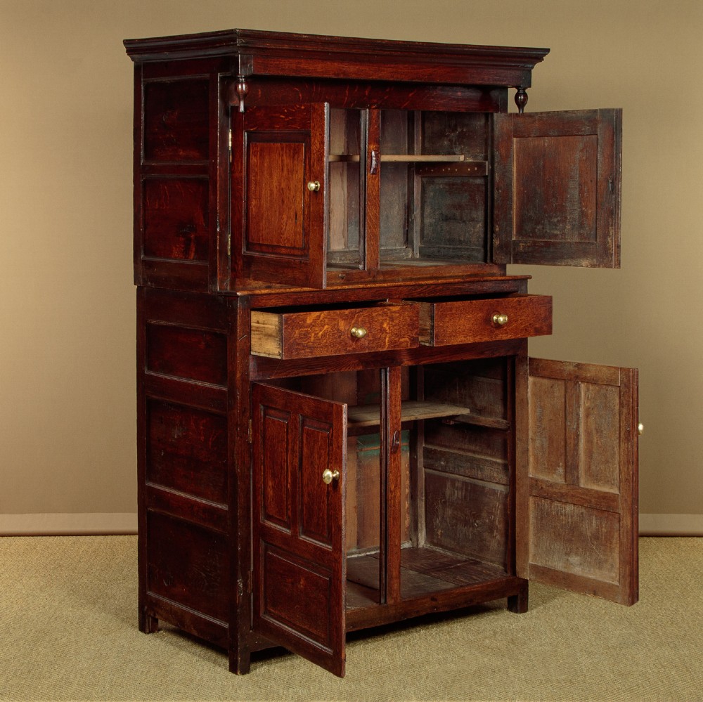 An 18th-century dark Welsh oak cwpwrdd deuddarn featuring an overhanging canopy with drop pendants, upper fielded panel doors, two drawers, and lower cupboard doors. View IV