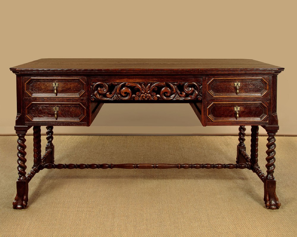 A dark carved oak writing desk featuring four geometric drawers, a scroll-carved central frieze, barley-twist legs, and a stretcher base.
