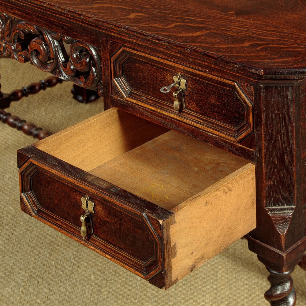 A dark carved oak writing desk featuring four geometric drawers, a scroll-carved central frieze, barley-twist legs, and a stretcher base. View IX