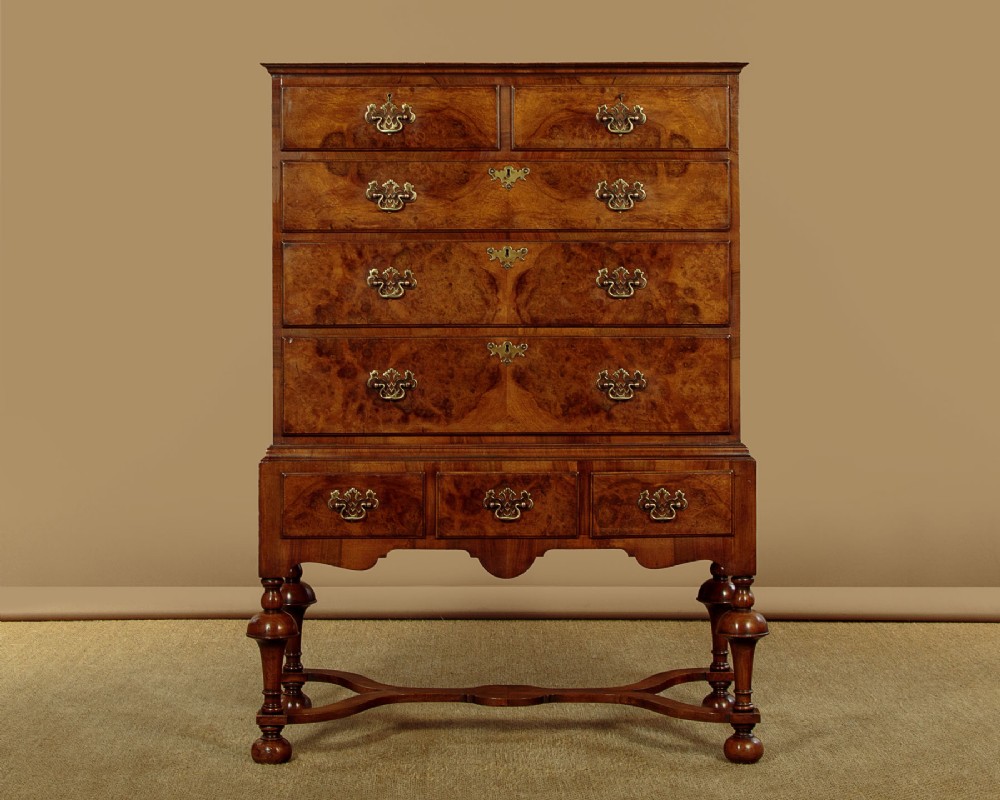 A 1930s Queen Anne Revival burr walnut chest on stand featuring multiple graduated drawers, decorative brass handles, turned legs, and a shaped apron.