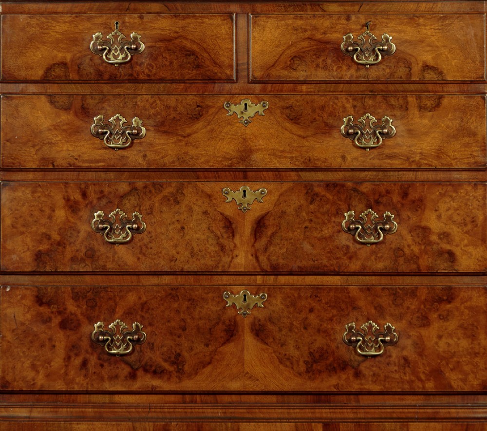 A 1930s Queen Anne Revival burr walnut chest on stand featuring multiple graduated drawers, decorative brass handles, turned legs, and a shaped apron. View VII
