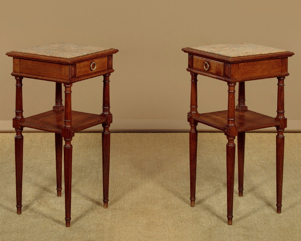 A pair of slender Edwardian oak lamp tables featuring inset marble tops, single drawers with brass ring pulls, turned tapered legs, and lower tier shelves.