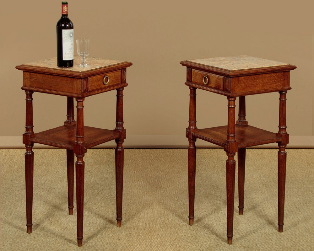 A pair of slender Edwardian oak lamp tables featuring inset marble tops, single drawers with brass ring pulls, turned tapered legs, and lower tier shelves. View II
