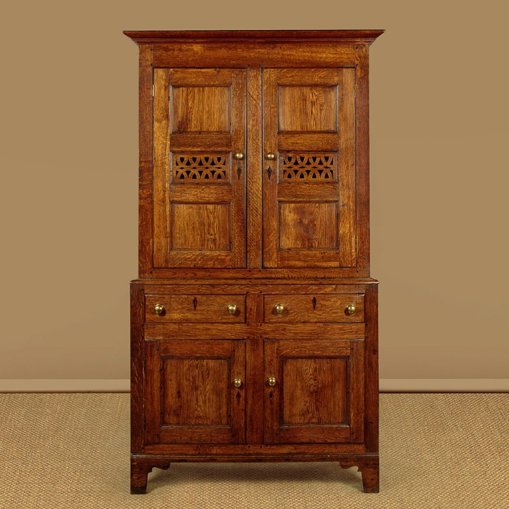 A two-part antique warm brown oak bread and cheese cupboard featuring upper doors with lattice ventilation fretwork over lower drawers and panelled cupboard doors.