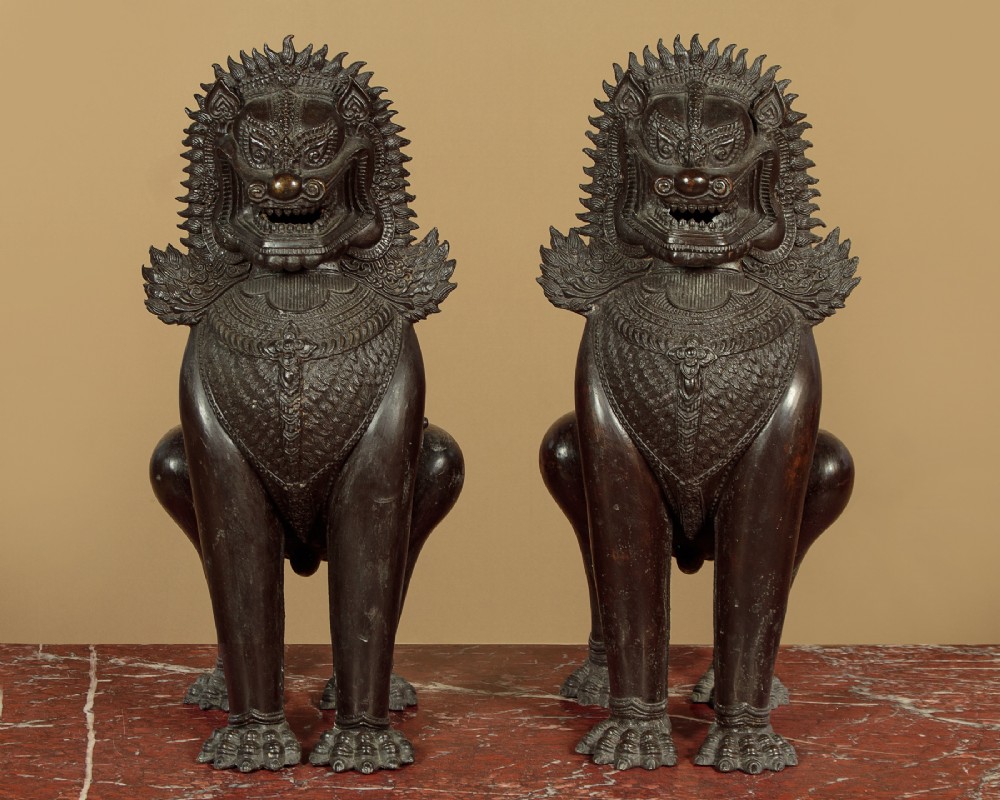 A pair of dark bronze temple lion statues sitting upright with intricate stylized manes and detailed ornamental chest bands. View III
