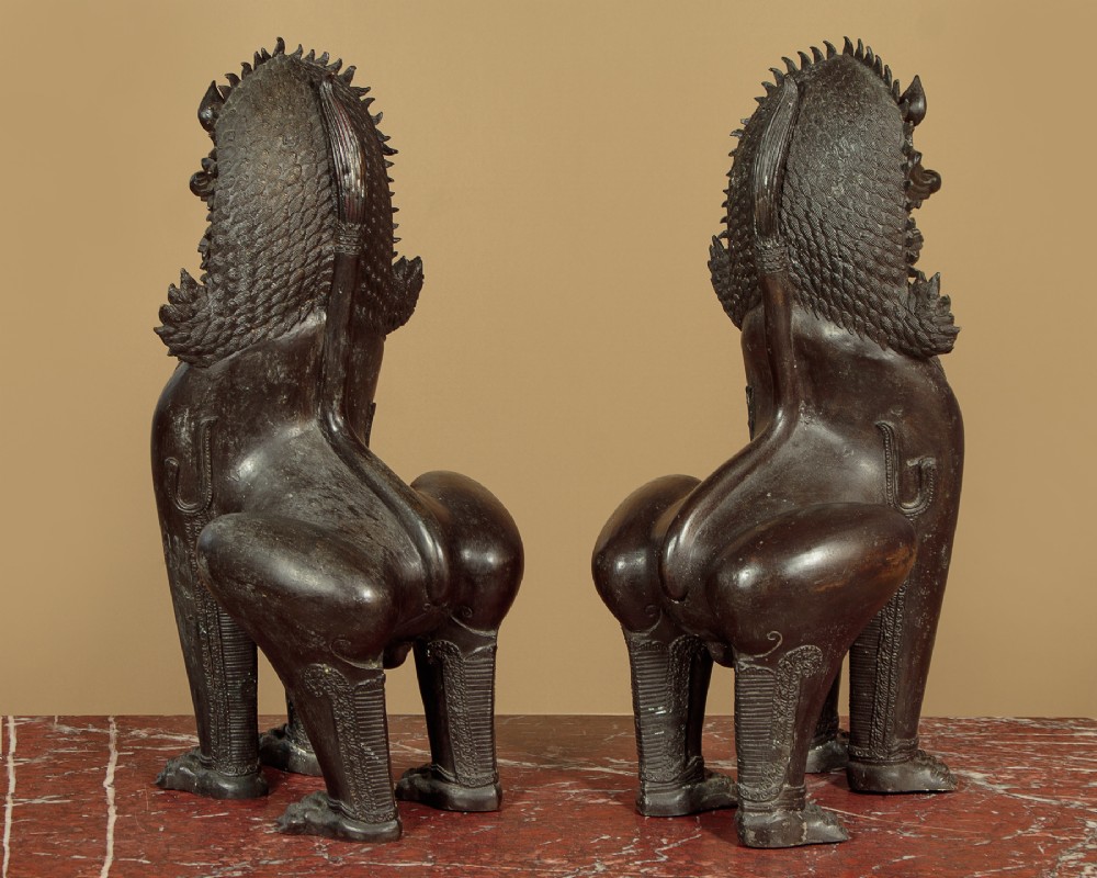 A pair of dark bronze temple lion statues sitting upright with intricate stylized manes and detailed ornamental chest bands. View IV