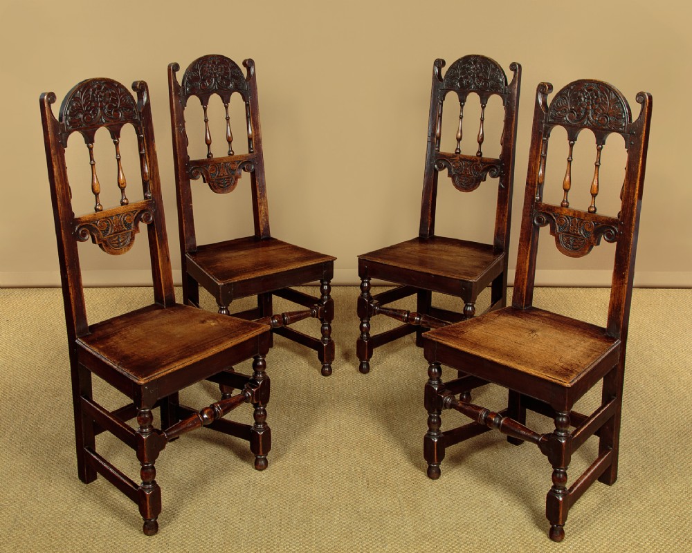 A set of four dark oak dining chairs featuring carved crest rails, arcaded spindle backs, wooden seats, and turned front legs.