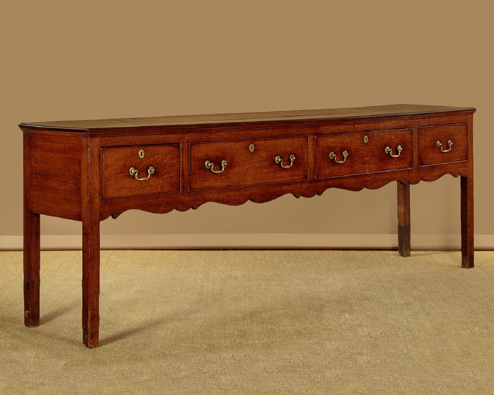 An 18th-century long oak dresser base featuring four drawers with brass swan-neck handles, a scalloped lower apron, and straight square legs.