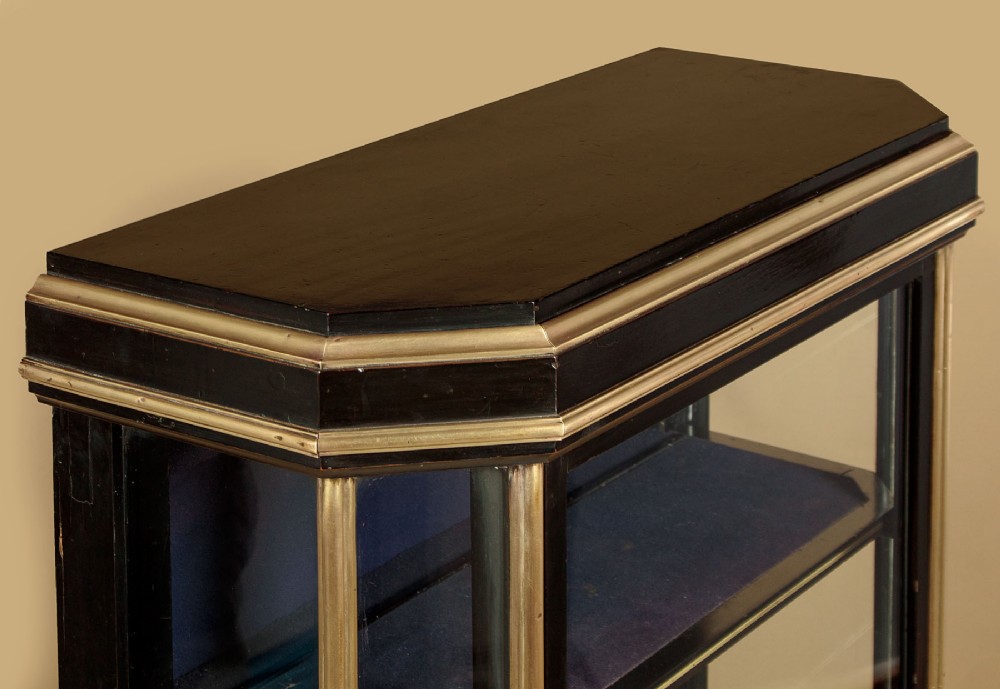 An Art Deco ebonised tall display cabinet featuring brass mounts, glazed side panels, a single glass door, and fabric-lined interior shelving. View VI