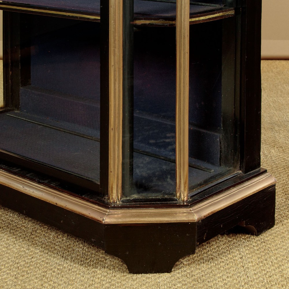 An Art Deco ebonised tall display cabinet featuring brass mounts, glazed side panels, a single glass door, and fabric-lined interior shelving. View XI