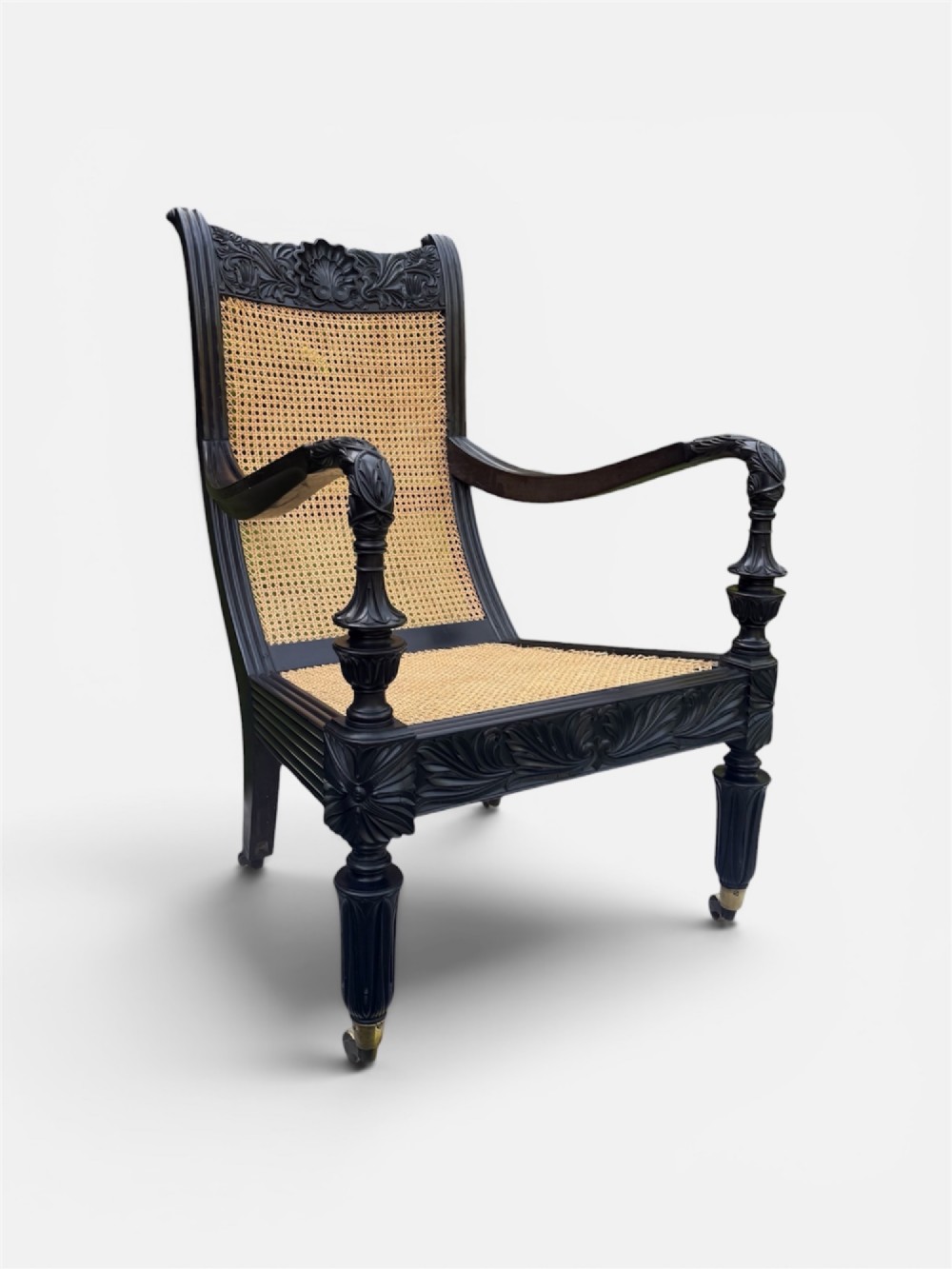 A mid-19th century carved ebony armchair featuring an intricately carved floral crest, woven natural cane seat and back, and turned fluted legs on brass castors.