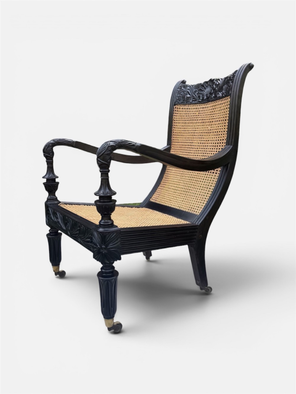 A mid-19th century carved ebony armchair featuring an intricately carved floral crest, woven natural cane seat and back, and turned fluted legs on brass castors. View II