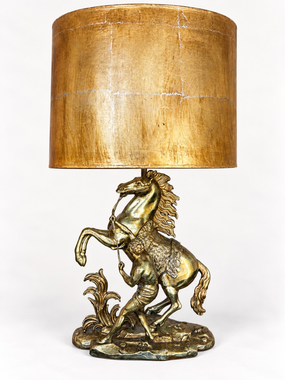 A sculptural brass table lamp depicting a rearing horse and groom after Guillaume Coustou, topped with a tall cylindrical gold leaf drum shade.