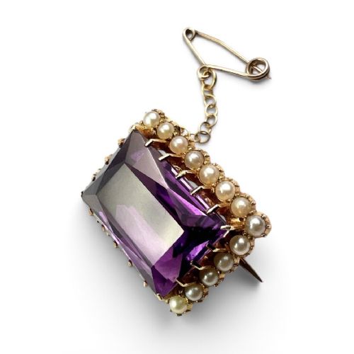 A rectangular Victorian brooch featuring a central facet-cut purple amethyst encircled by a halo of small white pearls set in 15ct gold with an attached safety chain. View II