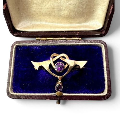 An Art Nouveau 9ct gold brooch featuring a heart-shaped whiplash frame set with a round purple amethyst and a suspended amethyst drop, displayed inside a fitted velvet case.