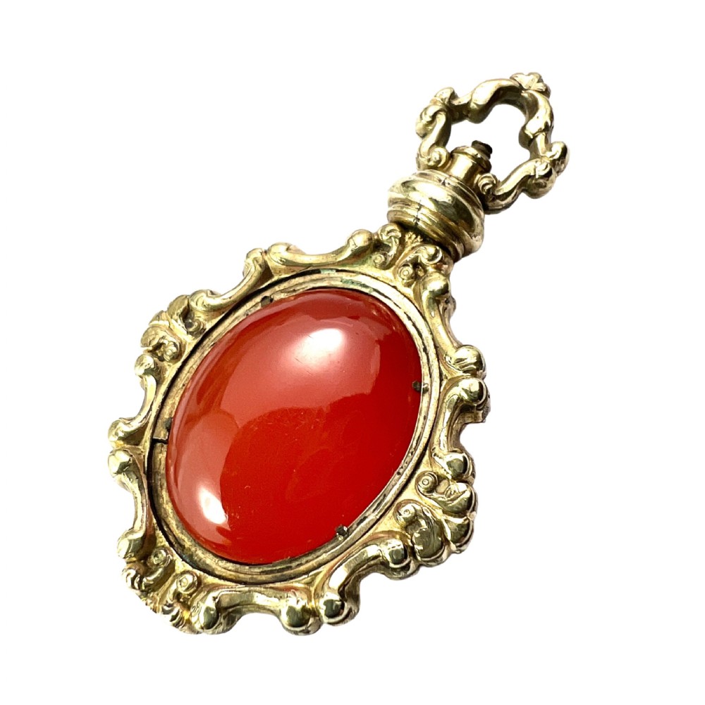 An ornate Georgian gilt metal swivel fob pendant featuring a polished oval red carnelian stone set within a scrollwork frame.