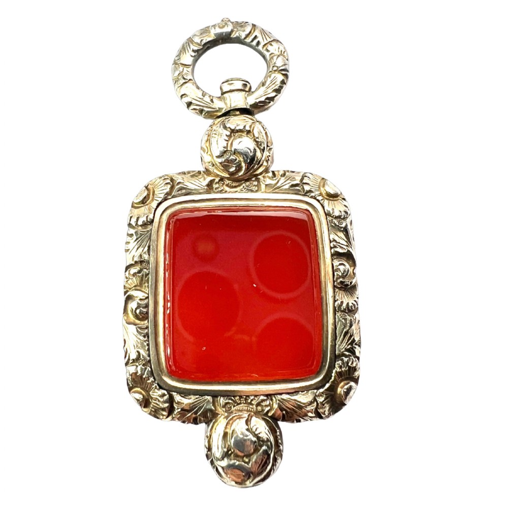 A Georgian era yellow gold watch fob pendant set with a smooth, square red carnelian stone and surrounded by ornate chased scrollwork with a top loop.