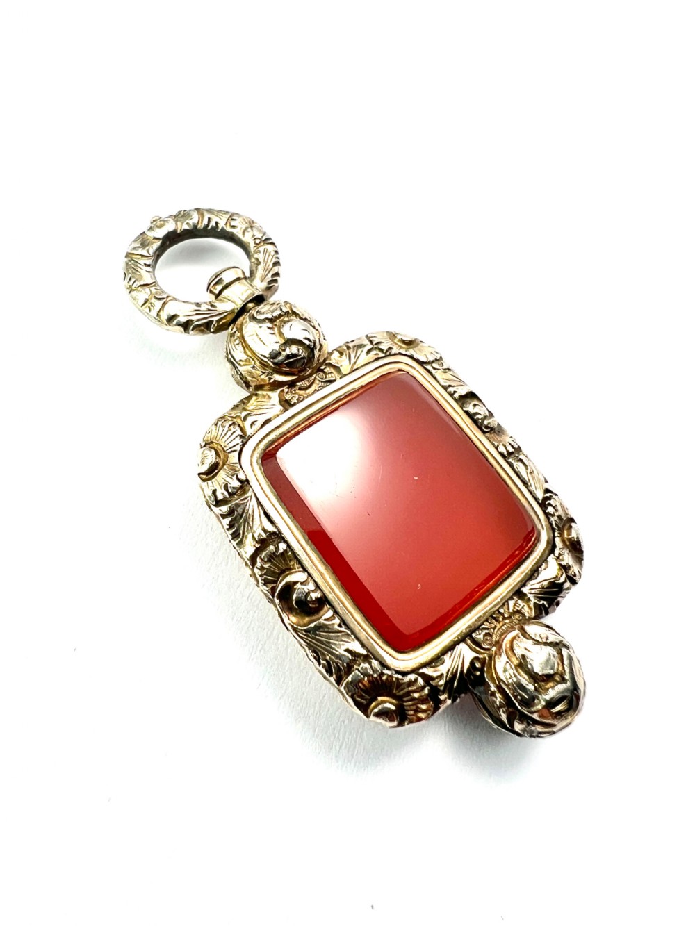 A Georgian era yellow gold watch fob pendant set with a smooth, square red carnelian stone and surrounded by ornate chased scrollwork with a top loop. View II