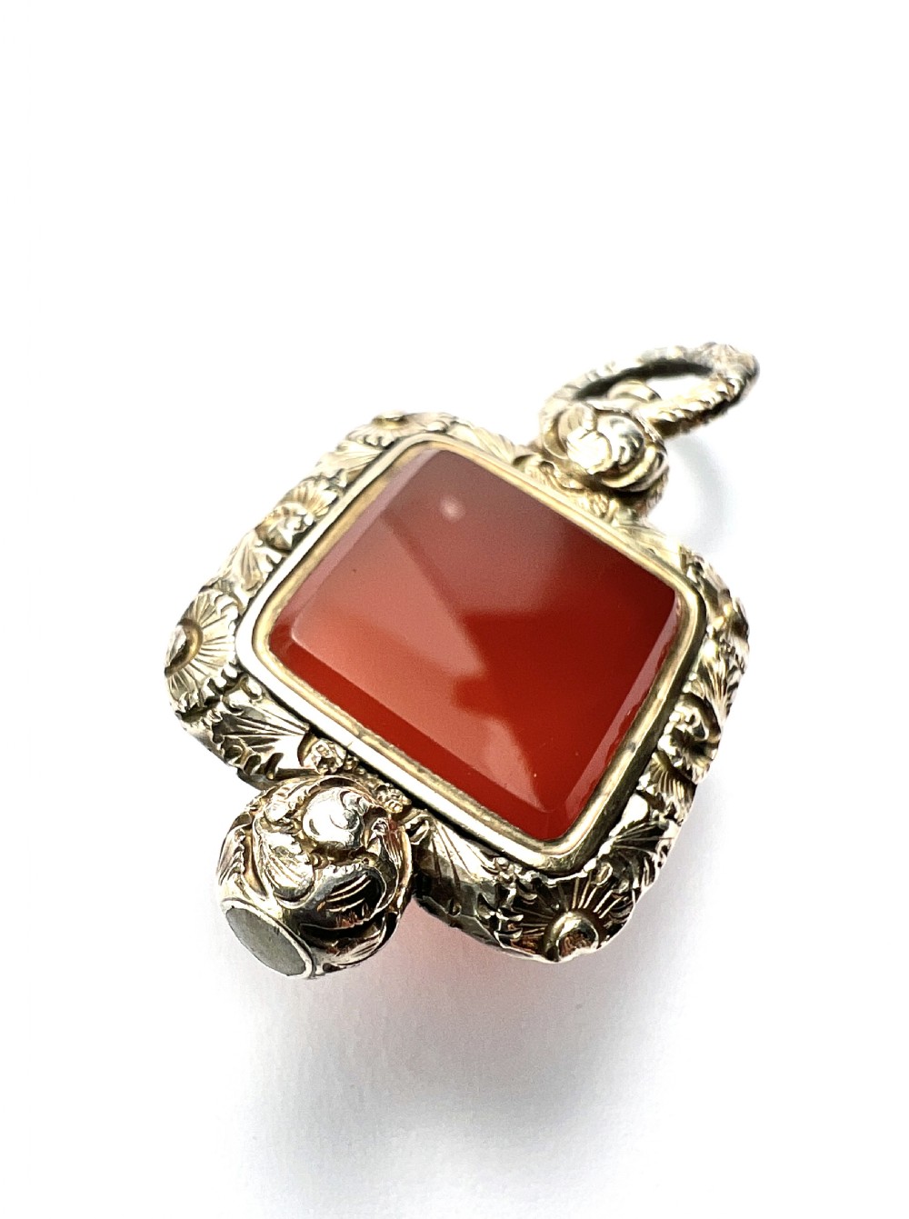 A Georgian era yellow gold watch fob pendant set with a smooth, square red carnelian stone and surrounded by ornate chased scrollwork with a top loop. View VI