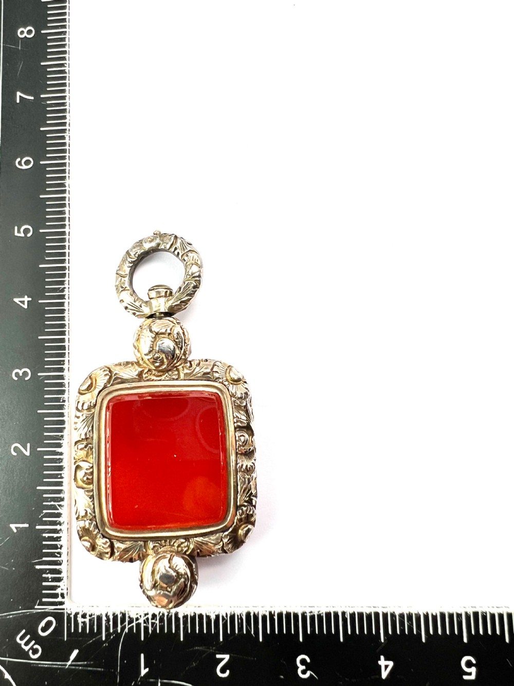 A Georgian era yellow gold watch fob pendant set with a smooth, square red carnelian stone and surrounded by ornate chased scrollwork with a top loop. View VII