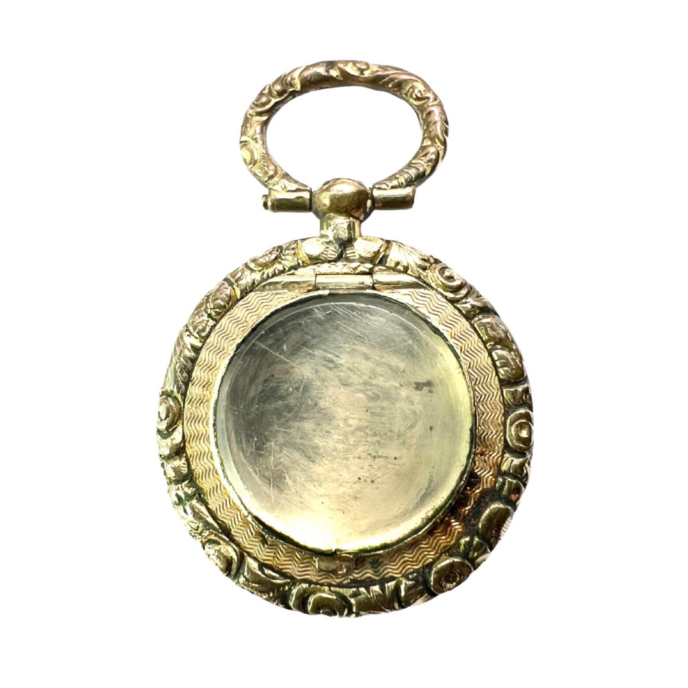 A circular antique gold mourning locket watch fob pendant featuring an intricately engraved foliate border, a textured ring, a glazed central compartment, and a top suspension loop.