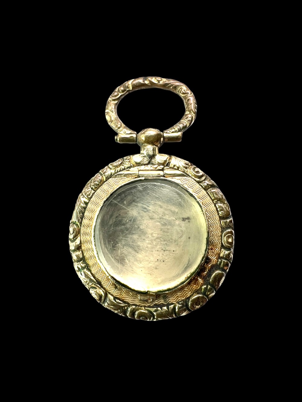 A circular antique gold mourning locket watch fob pendant featuring an intricately engraved foliate border, a textured ring, a glazed central compartment, and a top suspension loop. View II
