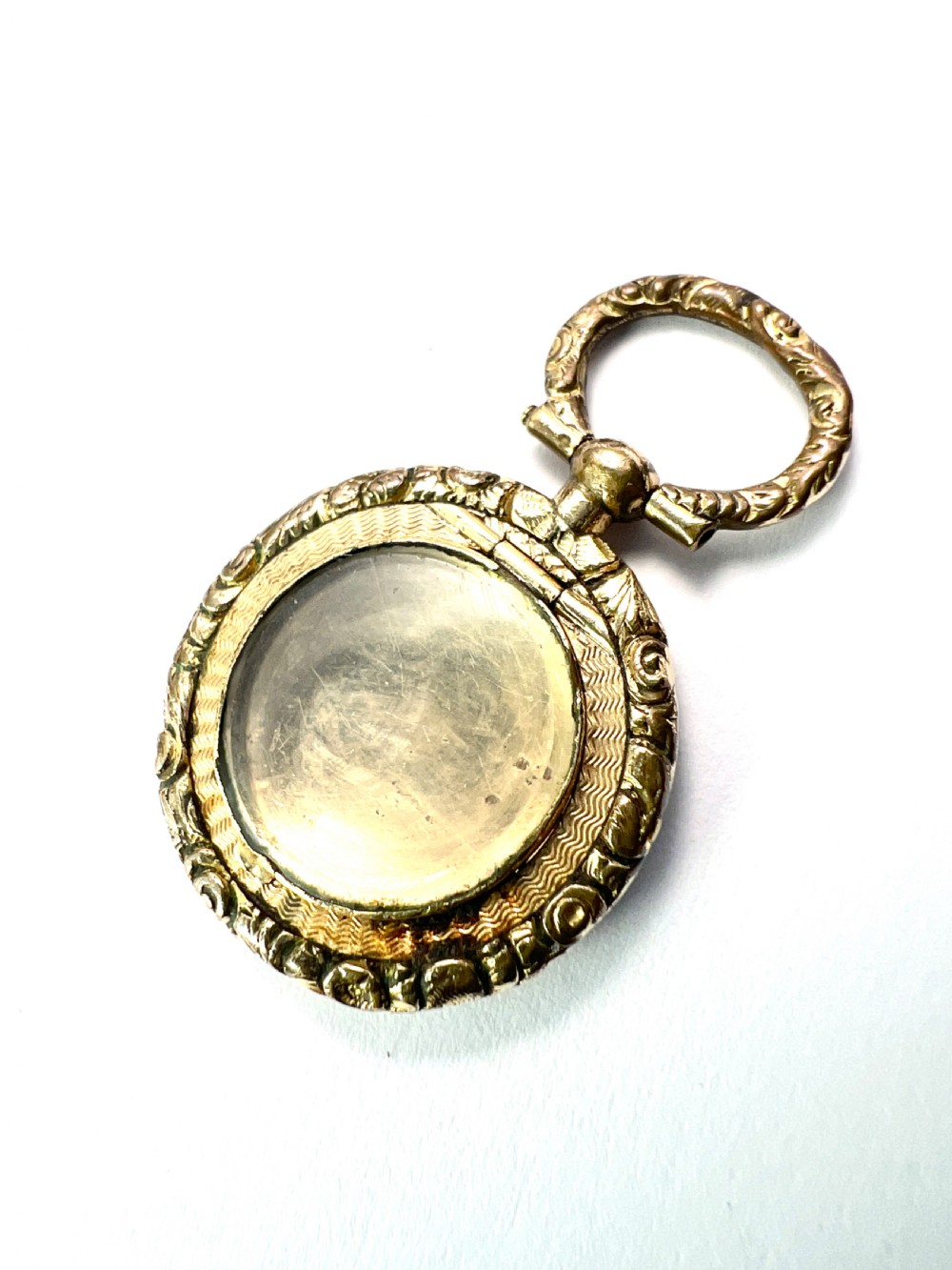 A circular antique gold mourning locket watch fob pendant featuring an intricately engraved foliate border, a textured ring, a glazed central compartment, and a top suspension loop. View III