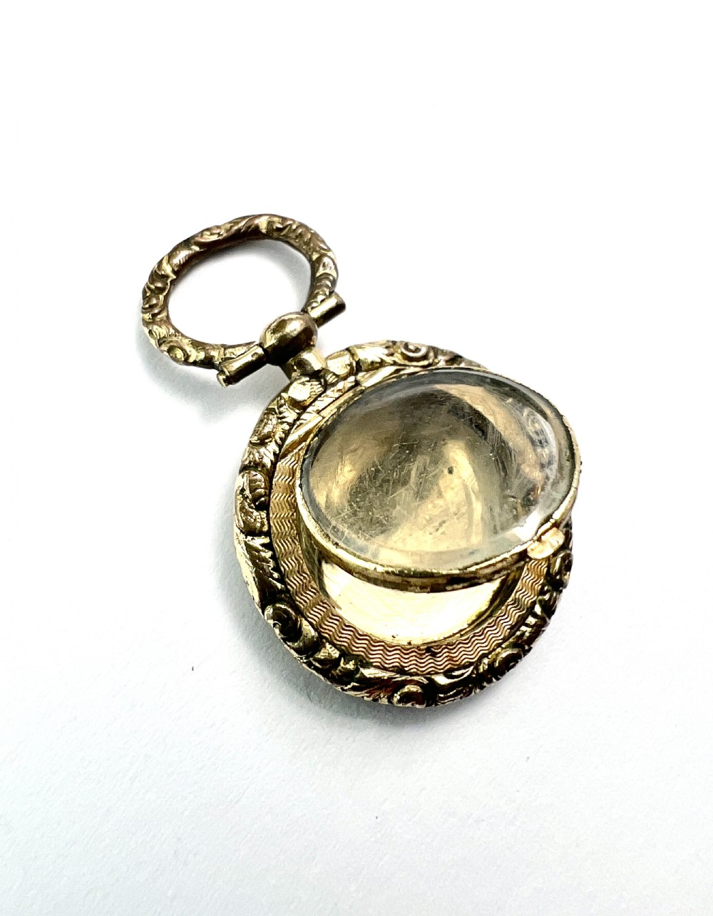 A circular antique gold mourning locket watch fob pendant featuring an intricately engraved foliate border, a textured ring, a glazed central compartment, and a top suspension loop. View V