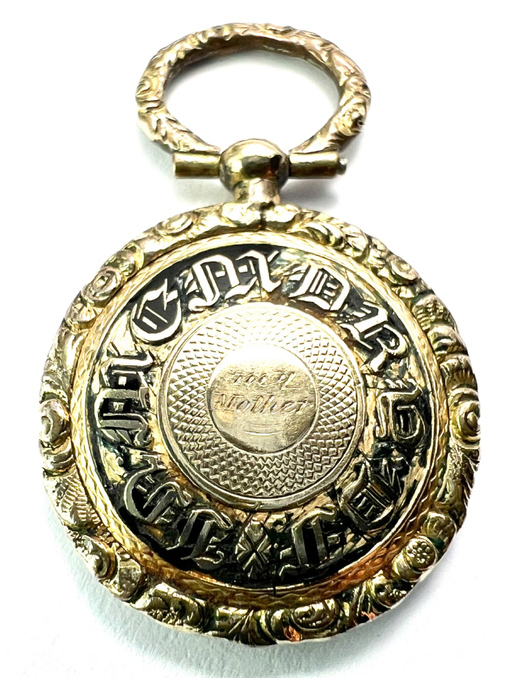 A circular antique gold mourning locket watch fob pendant featuring an intricately engraved foliate border, a textured ring, a glazed central compartment, and a top suspension loop. View VI