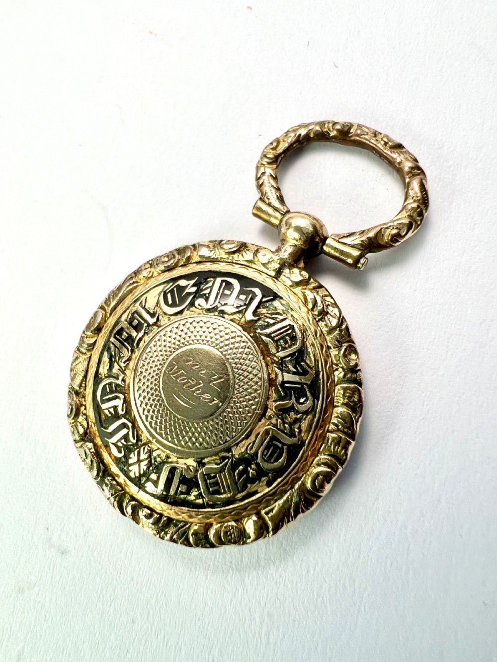 A circular antique gold mourning locket watch fob pendant featuring an intricately engraved foliate border, a textured ring, a glazed central compartment, and a top suspension loop. View VII