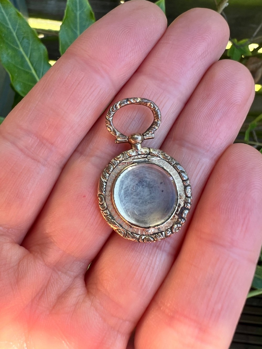 A circular antique gold mourning locket watch fob pendant featuring an intricately engraved foliate border, a textured ring, a glazed central compartment, and a top suspension loop. View VIII