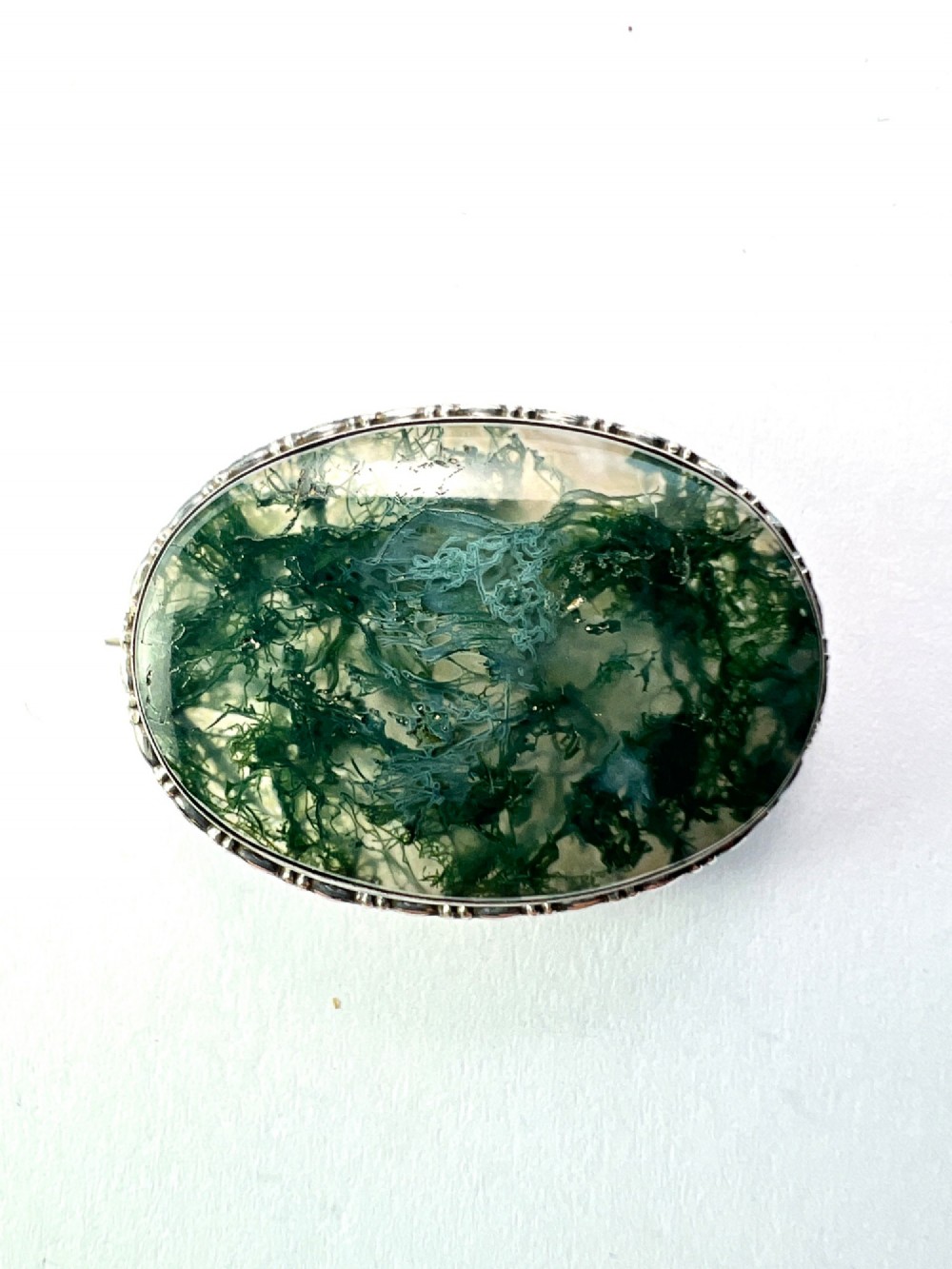 An oval antique sterling silver brooch featuring a translucent moss agate cabochon with green dendritic inclusions within an engraved bezel frame.