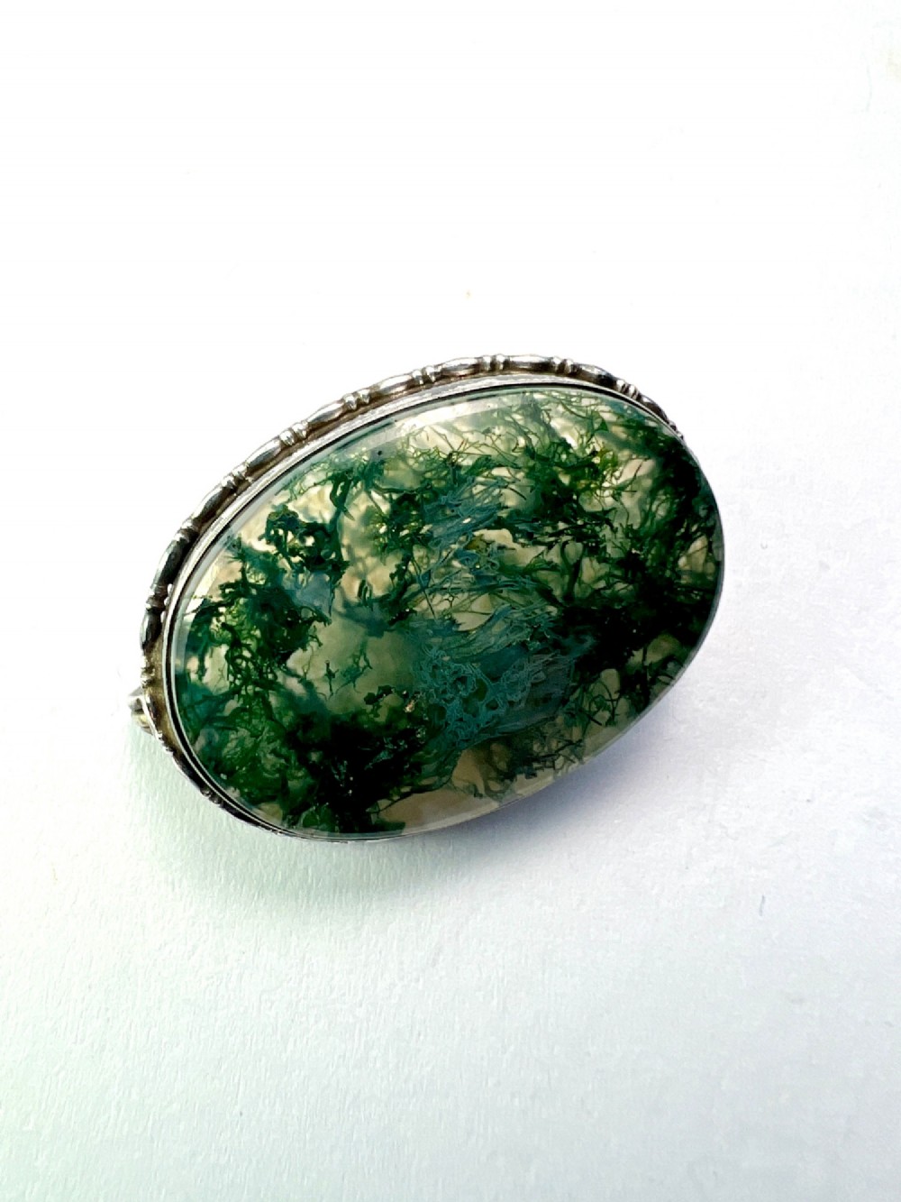 An oval antique sterling silver brooch featuring a translucent moss agate cabochon with green dendritic inclusions within an engraved bezel frame. View III