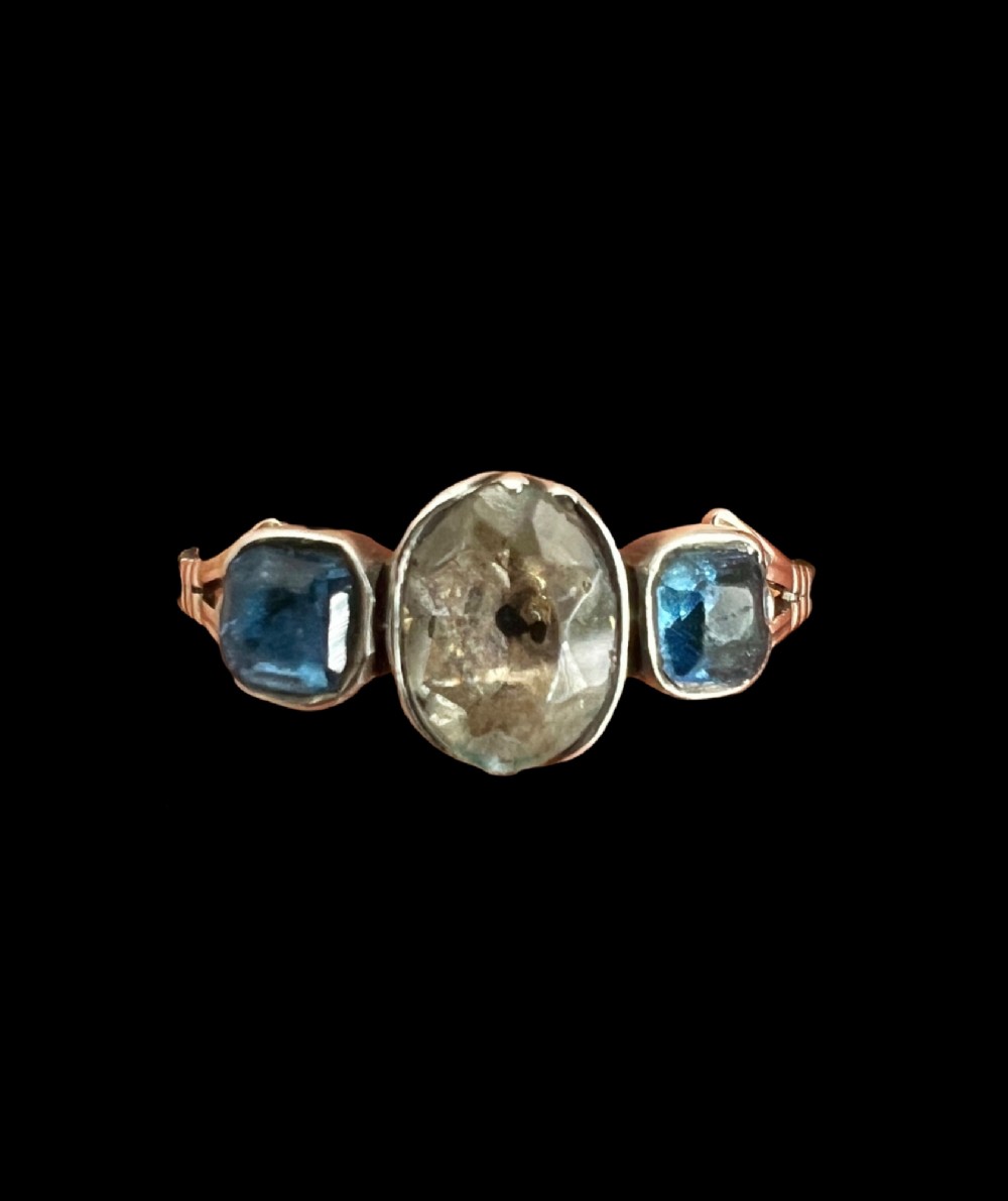 A Georgian three-stone ring featuring a central oval clear paste stone flanked by two blue cushion-cut paste stones set in silver and gold. View II