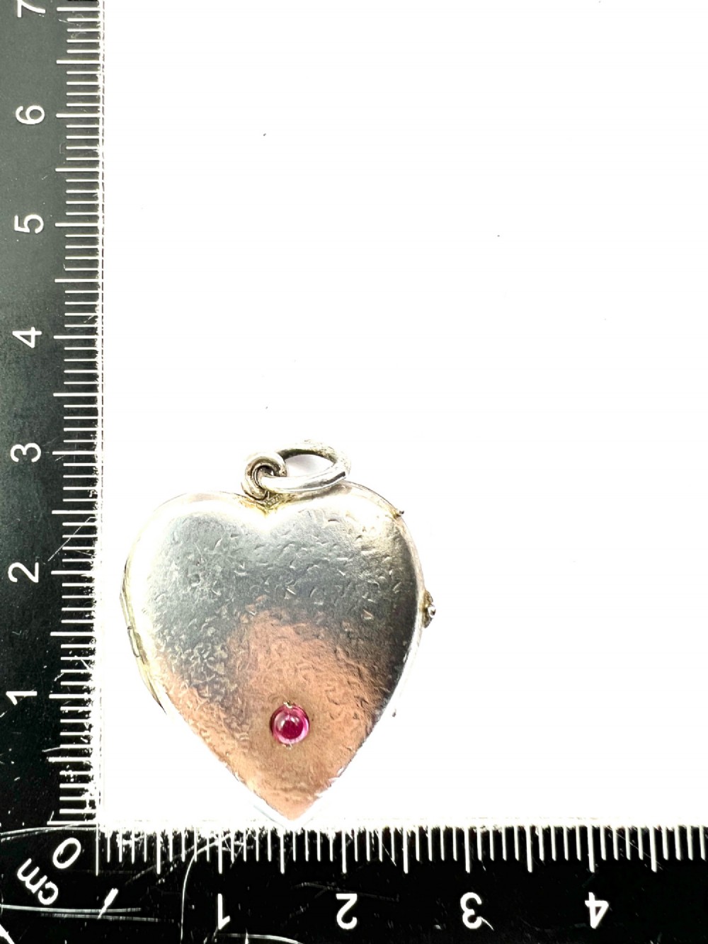 A textured silver heart-shaped locket pendant with a flush-set red cabochon stone and a top bail ring. View VI