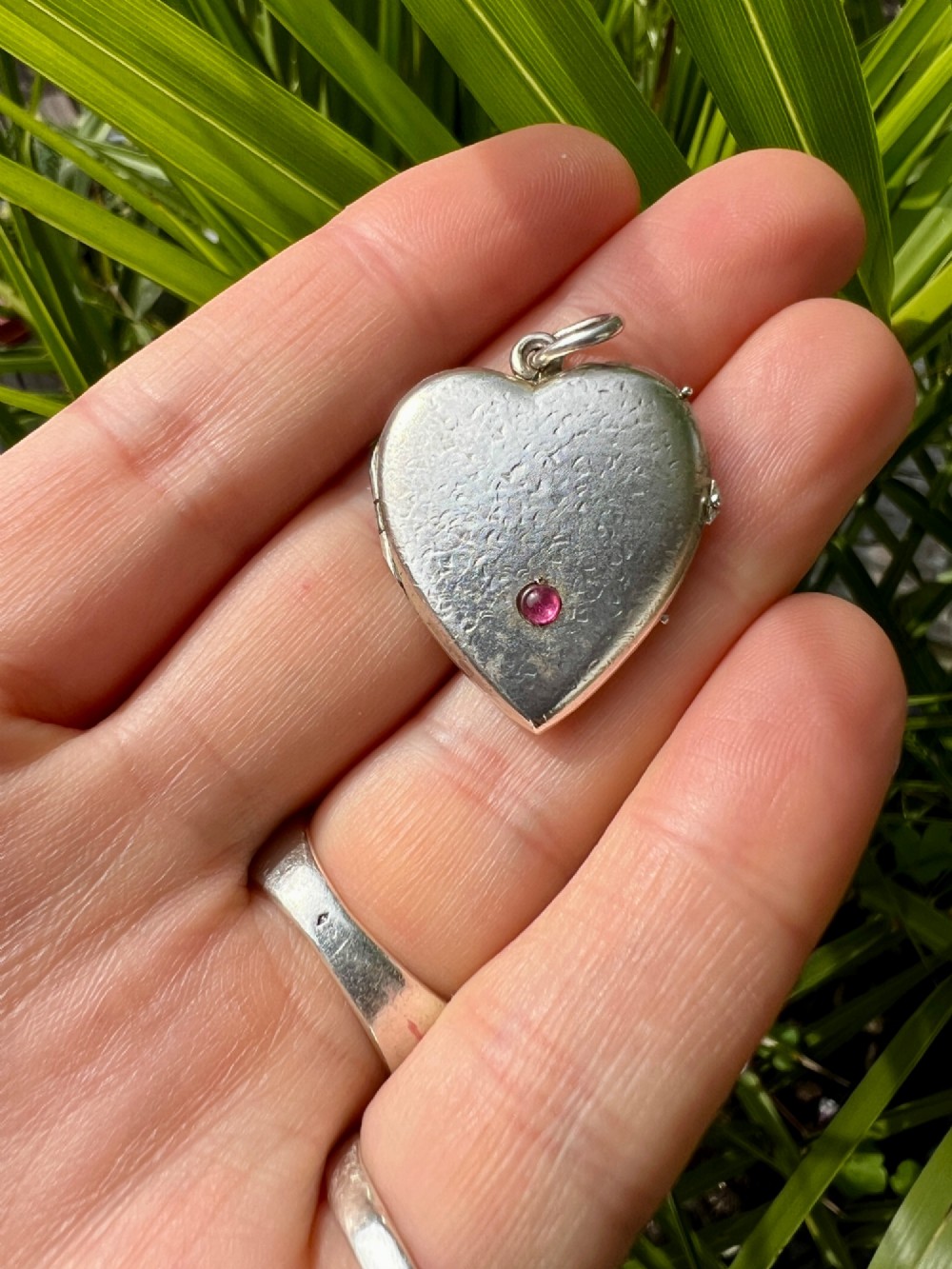 A textured silver heart-shaped locket pendant with a flush-set red cabochon stone and a top bail ring. View VII