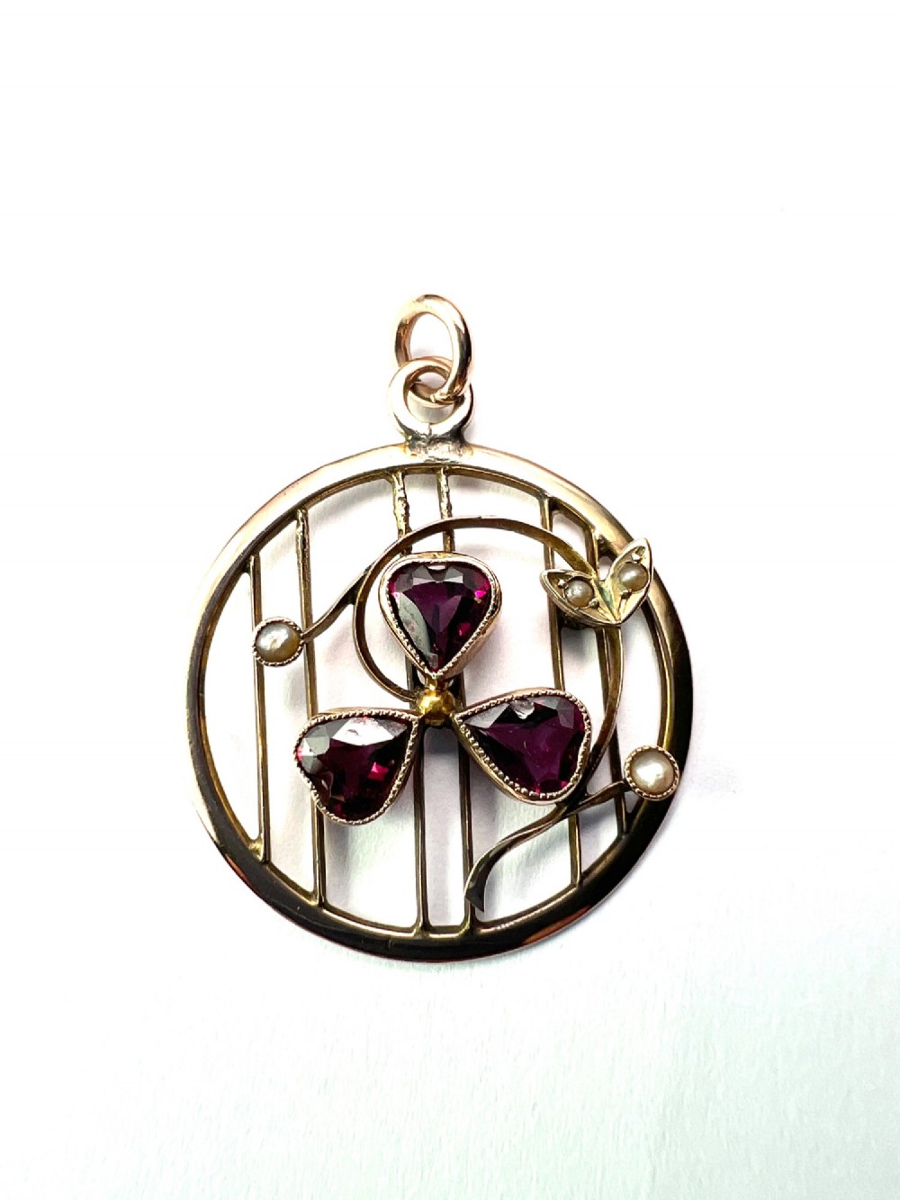 A circular 9ct gold Edwardian openwork pendant featuring three heart-cut deep red garnets forming a clover motif flanked by seed pearls. View VI