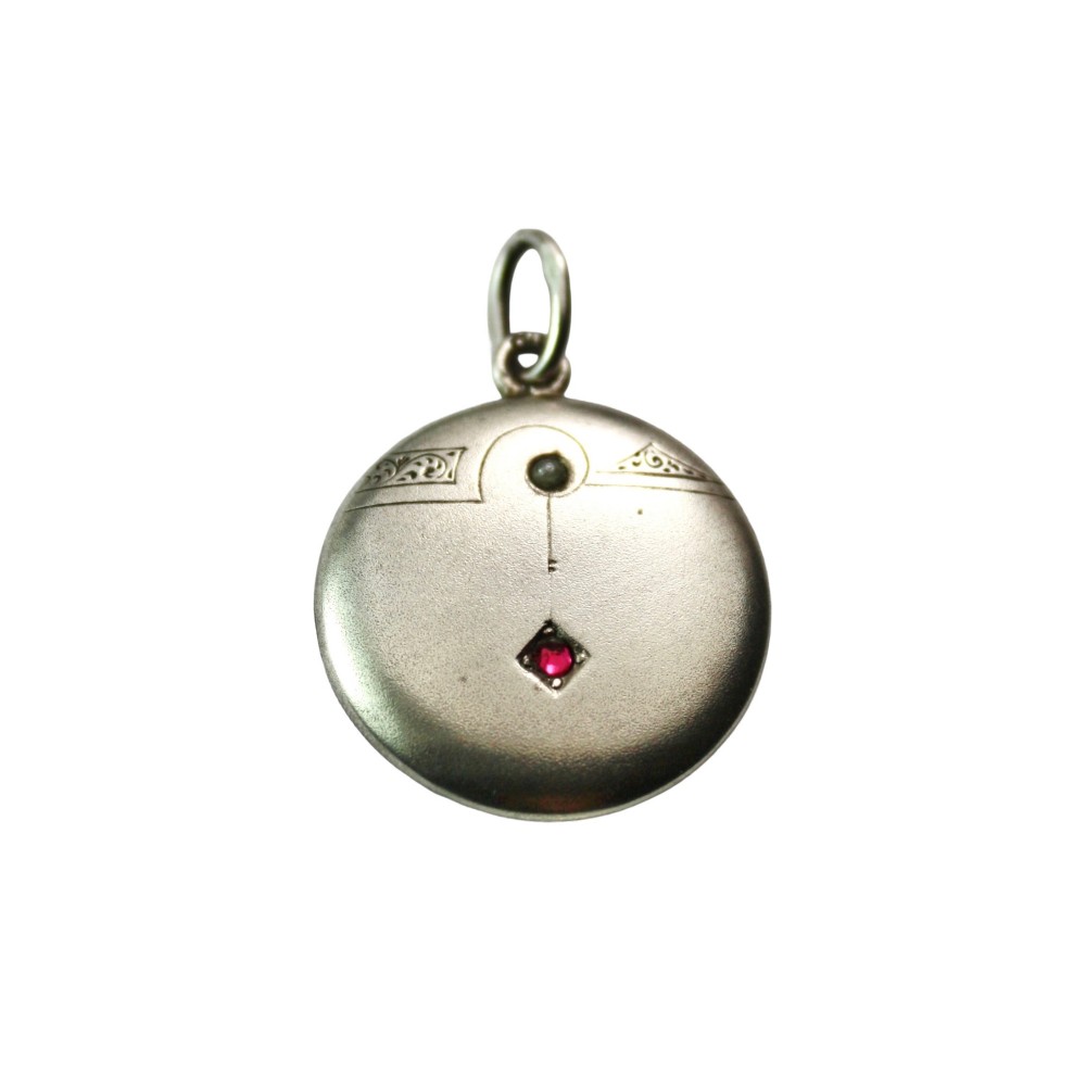 A circular silver Art Nouveau pendant featuring engraved line details, a central seed pearl, and a flush-set red paste stone.