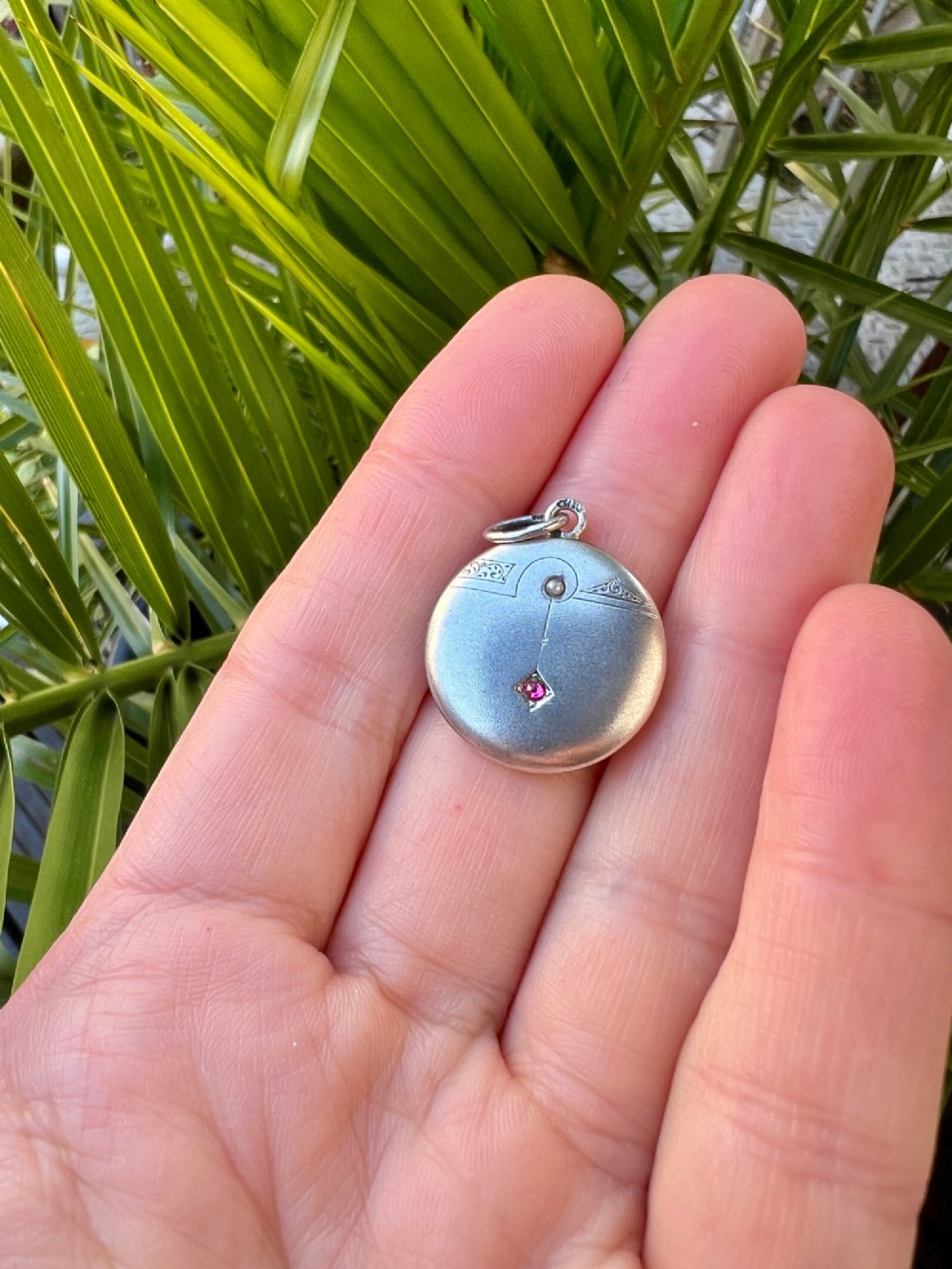 A circular silver Art Nouveau pendant featuring engraved line details, a central seed pearl, and a flush-set red paste stone. View IV
