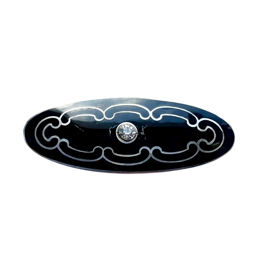 An oval Art Deco brooch featuring black enamel inlay with silver scroll linework and a central clear glass paste stone.