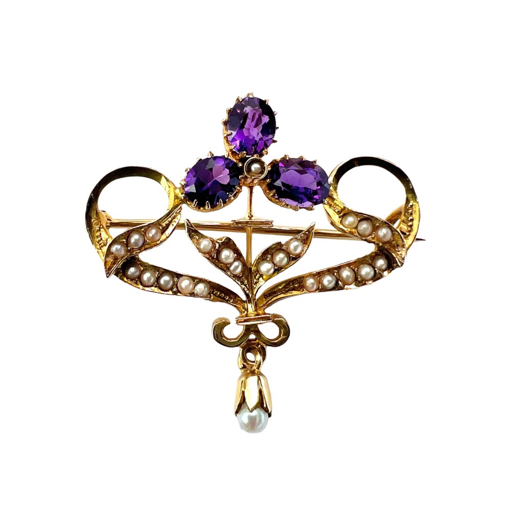 An Edwardian 9ct gold openwork brooch set with three oval-cut purple amethysts, seed pearls along scrolling ribbon motifs, and an articulated pearl drop.