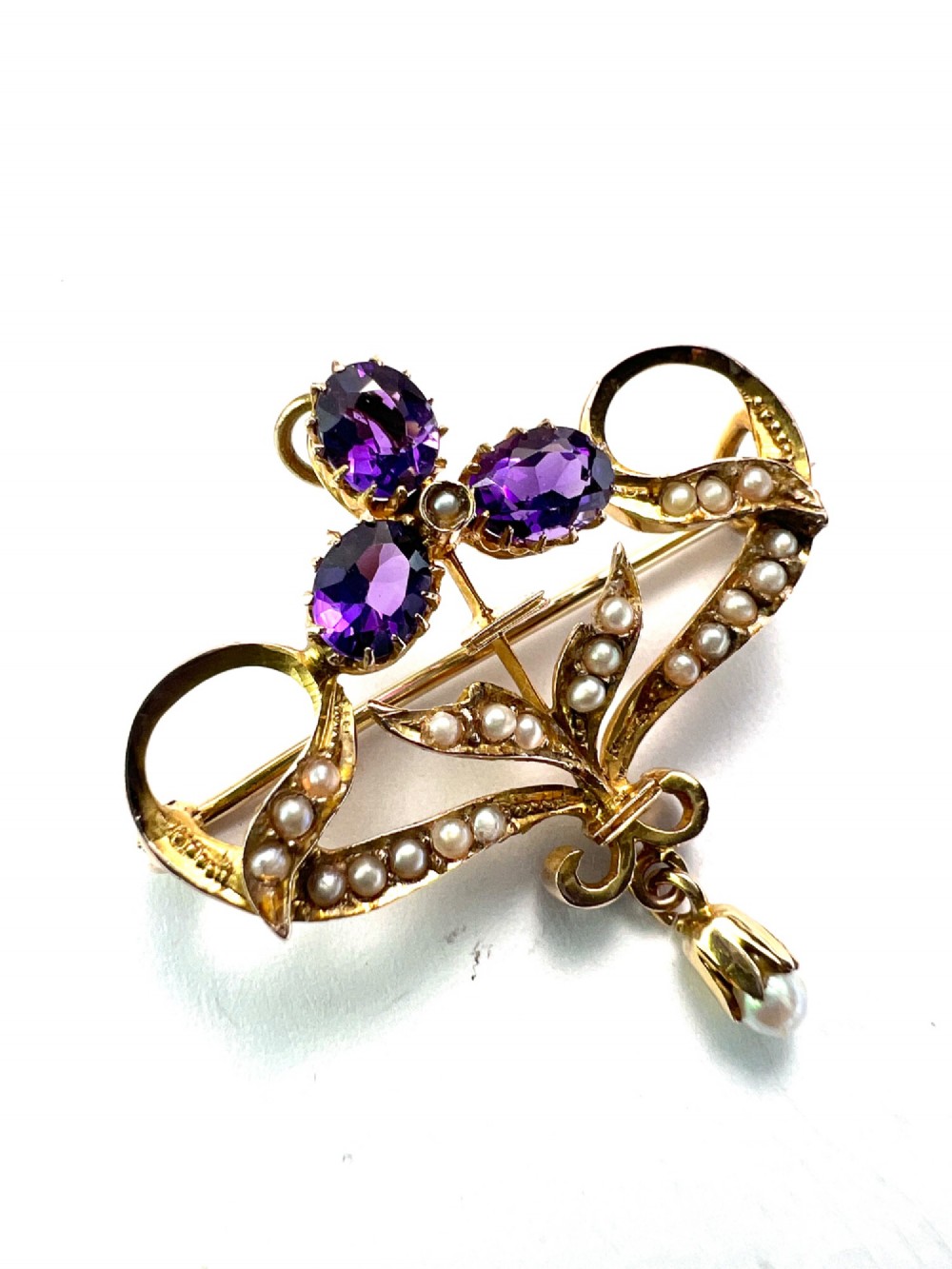 An Edwardian 9ct gold openwork brooch set with three oval-cut purple amethysts, seed pearls along scrolling ribbon motifs, and an articulated pearl drop. View II