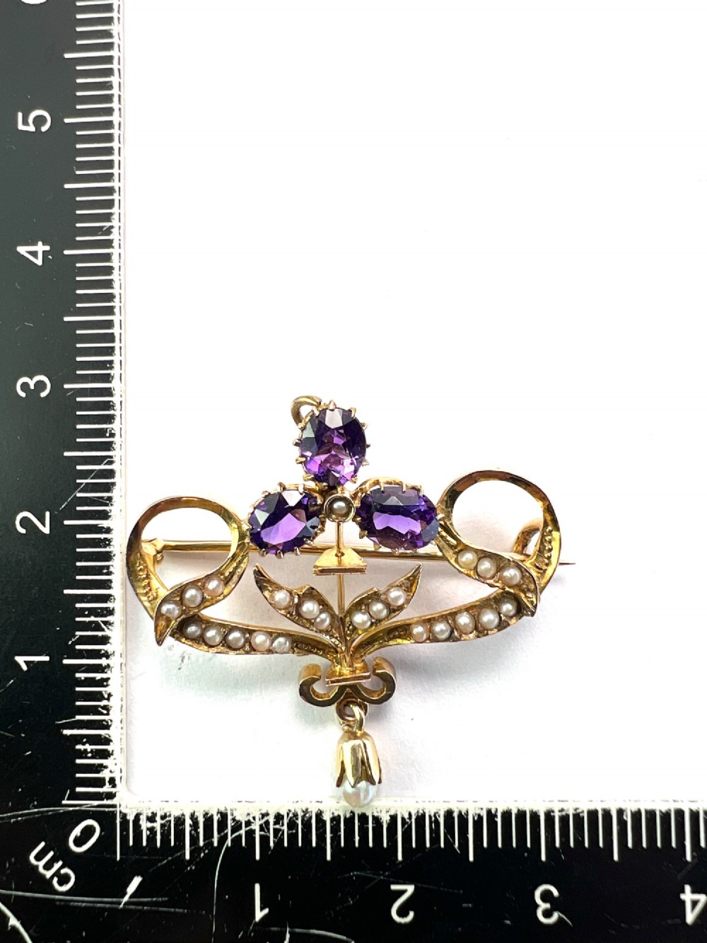 An Edwardian 9ct gold openwork brooch set with three oval-cut purple amethysts, seed pearls along scrolling ribbon motifs, and an articulated pearl drop. View IV