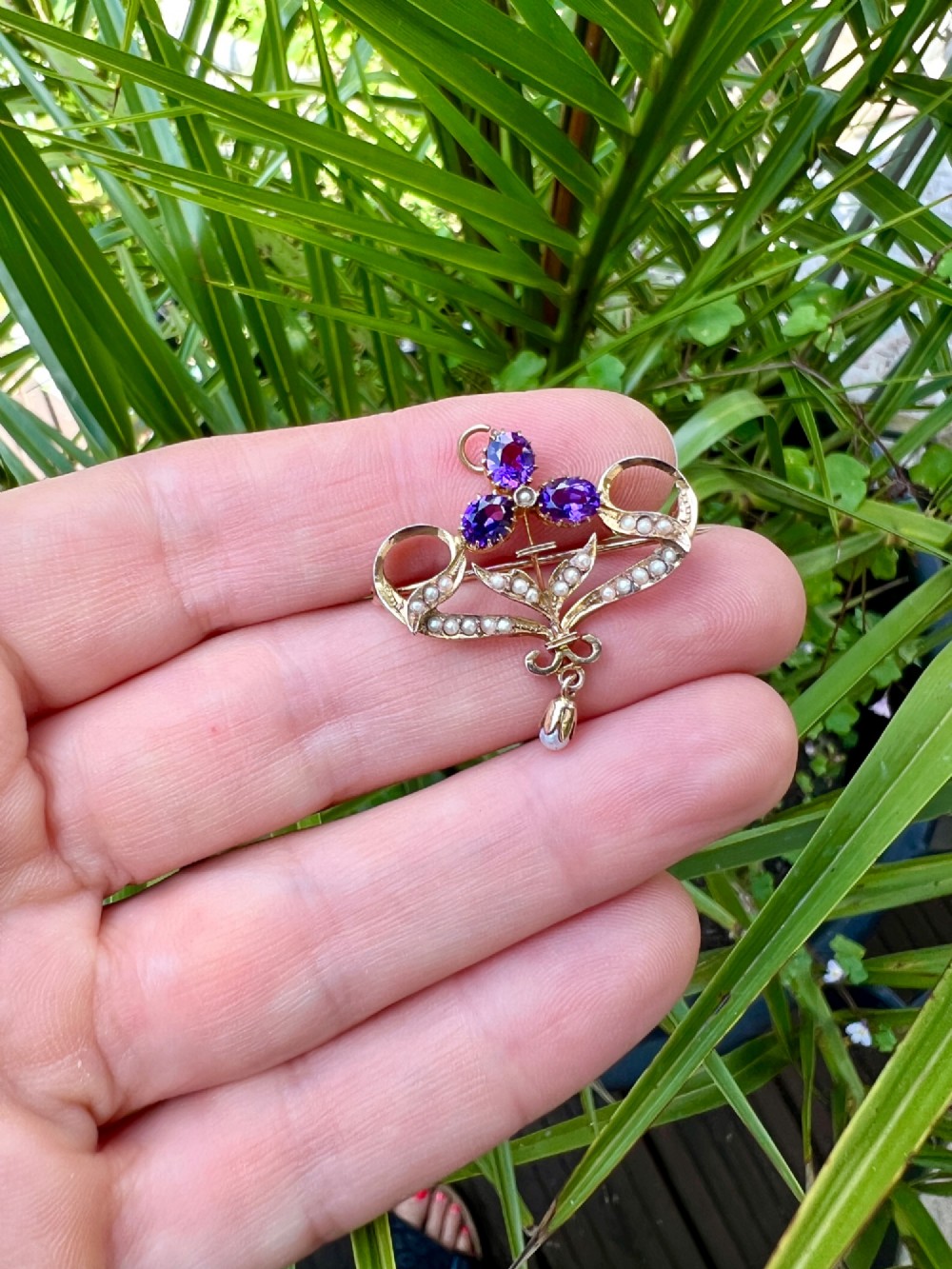 An Edwardian 9ct gold openwork brooch set with three oval-cut purple amethysts, seed pearls along scrolling ribbon motifs, and an articulated pearl drop. View V