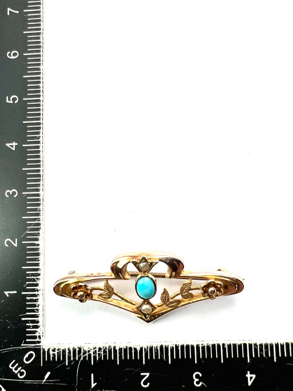 An Edwardian 9ct gold openwork bar brooch featuring a central oval cabochon turquoise flanked by two seed pearls, engraved leaves, and two small gold flowers. View IV