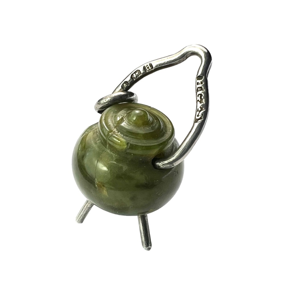 An Edwardian sterling silver and carved green stone cauldron pendant featuring tripod feet, a moveable silver handle, and hallmarked stamp details.