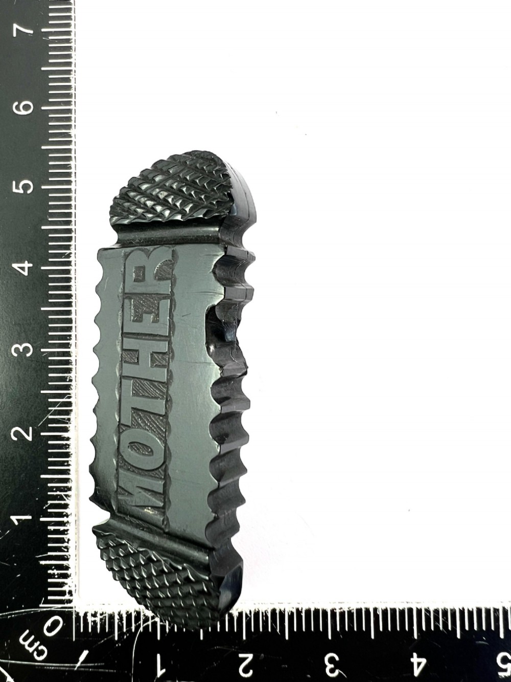 An oblong carved black Whitby jet brooch featuring the raised carved word MOTHER flanked by cross-hatched ends and notched borders. View V