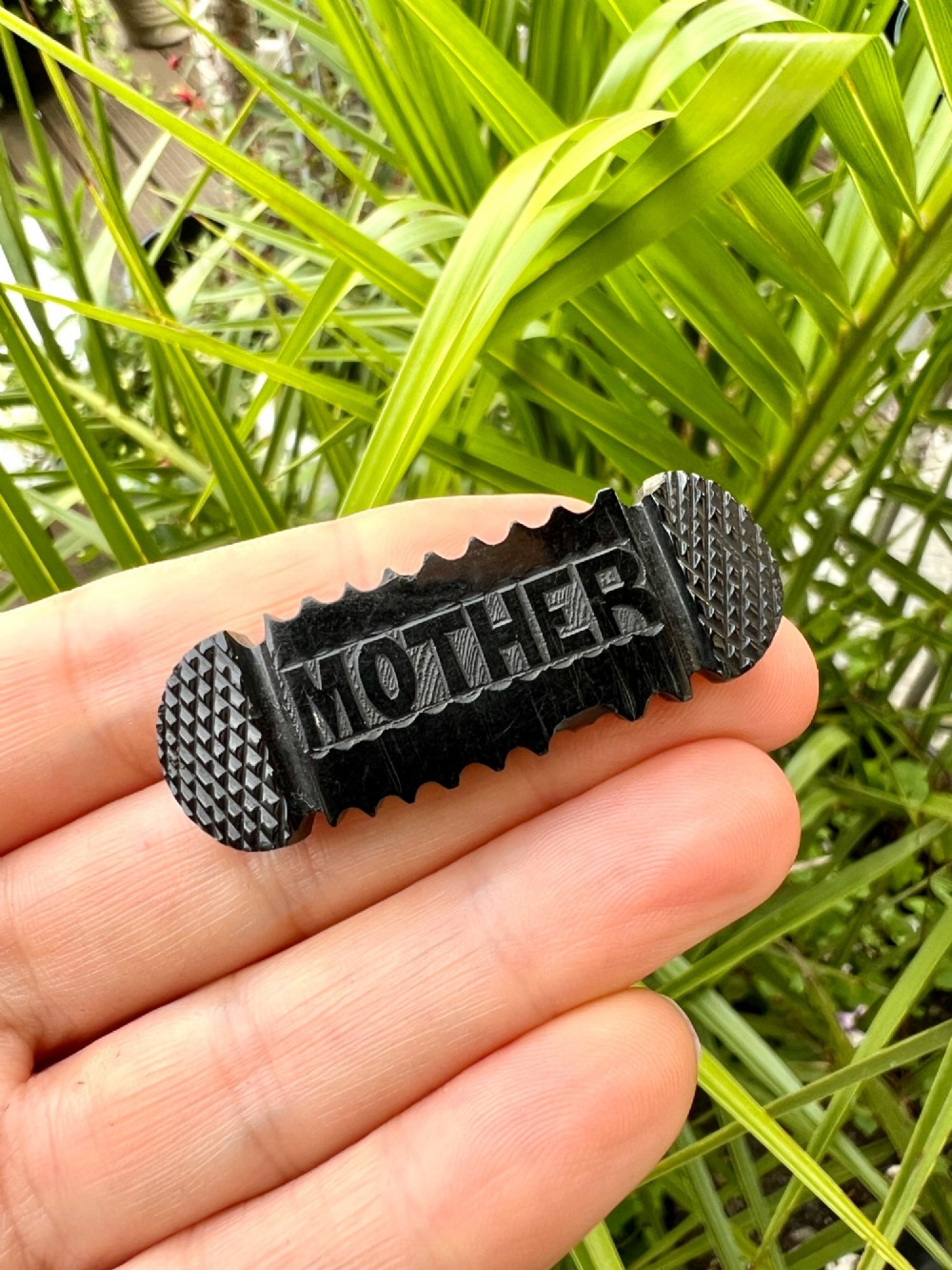 An oblong carved black Whitby jet brooch featuring the raised carved word MOTHER flanked by cross-hatched ends and notched borders. View VI